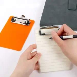 Orange Clipboard with notepad and pen with 2 hands writing on the notepad.